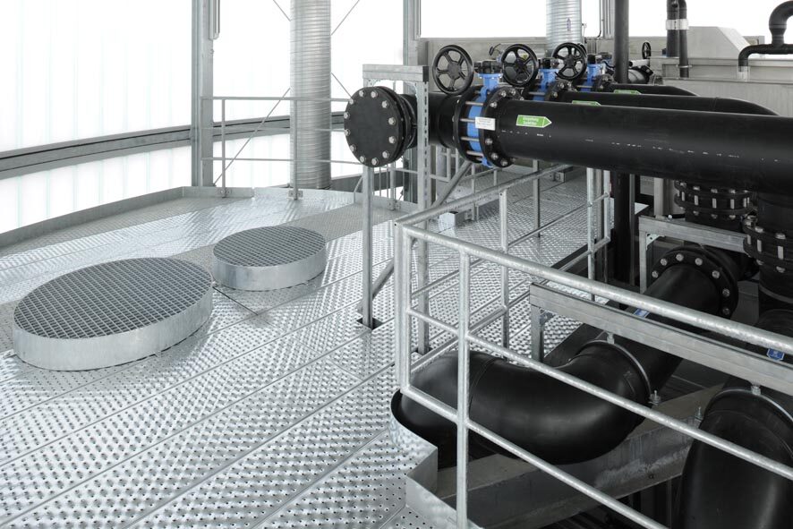 Innenbereich eines Pumphauses. Die schwarze Pumpe ist durch ein verzinktes Geländer gesichert. Die Plattform besteht aus verzinkten Blechprofilrosten mit runden nach oben gewölbten Luftschächten aus Gitterrosten.